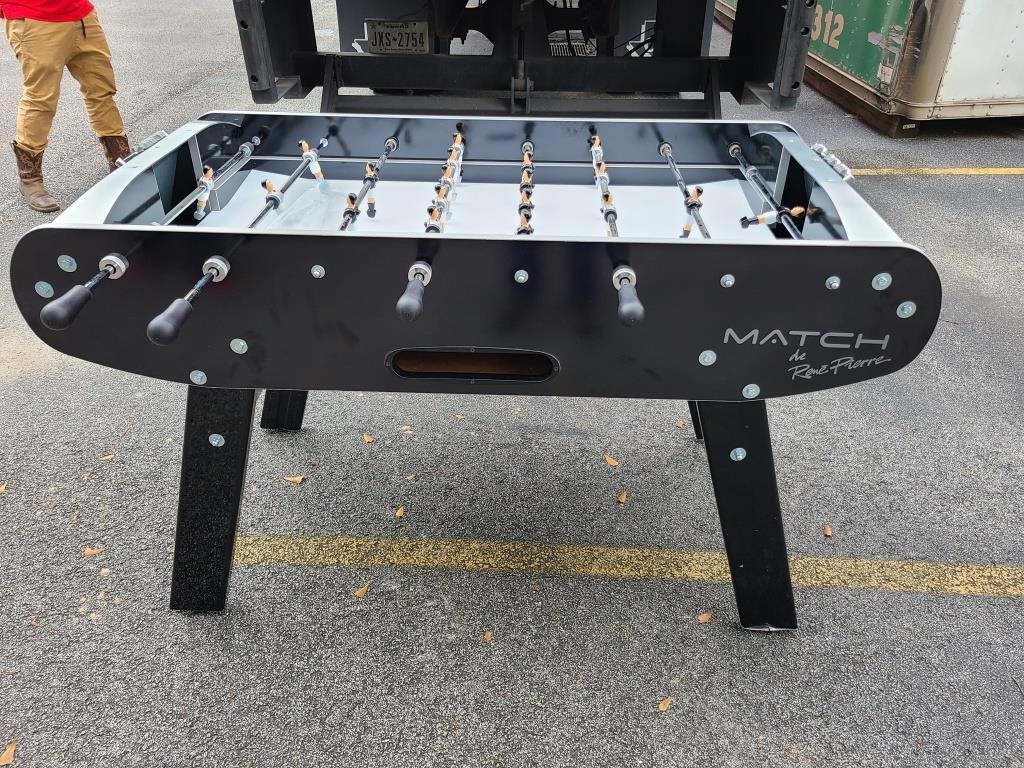 Rene Pierre Match Foosball Table in Black stands on asphalt with a person in boots nearby.