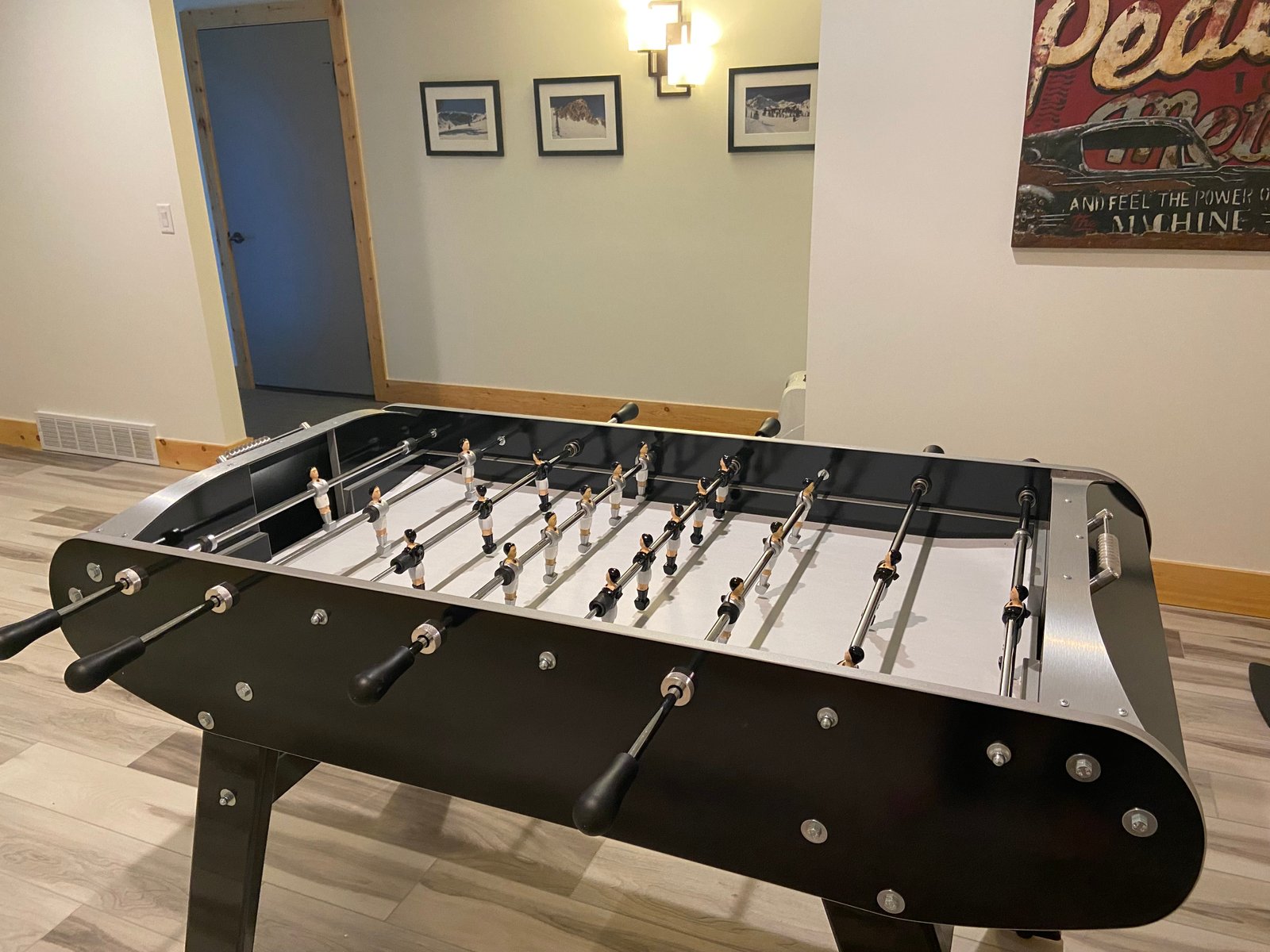 A Rene Pierre Match Foosball Table in black sits in a room with framed pictures and a retro sign.