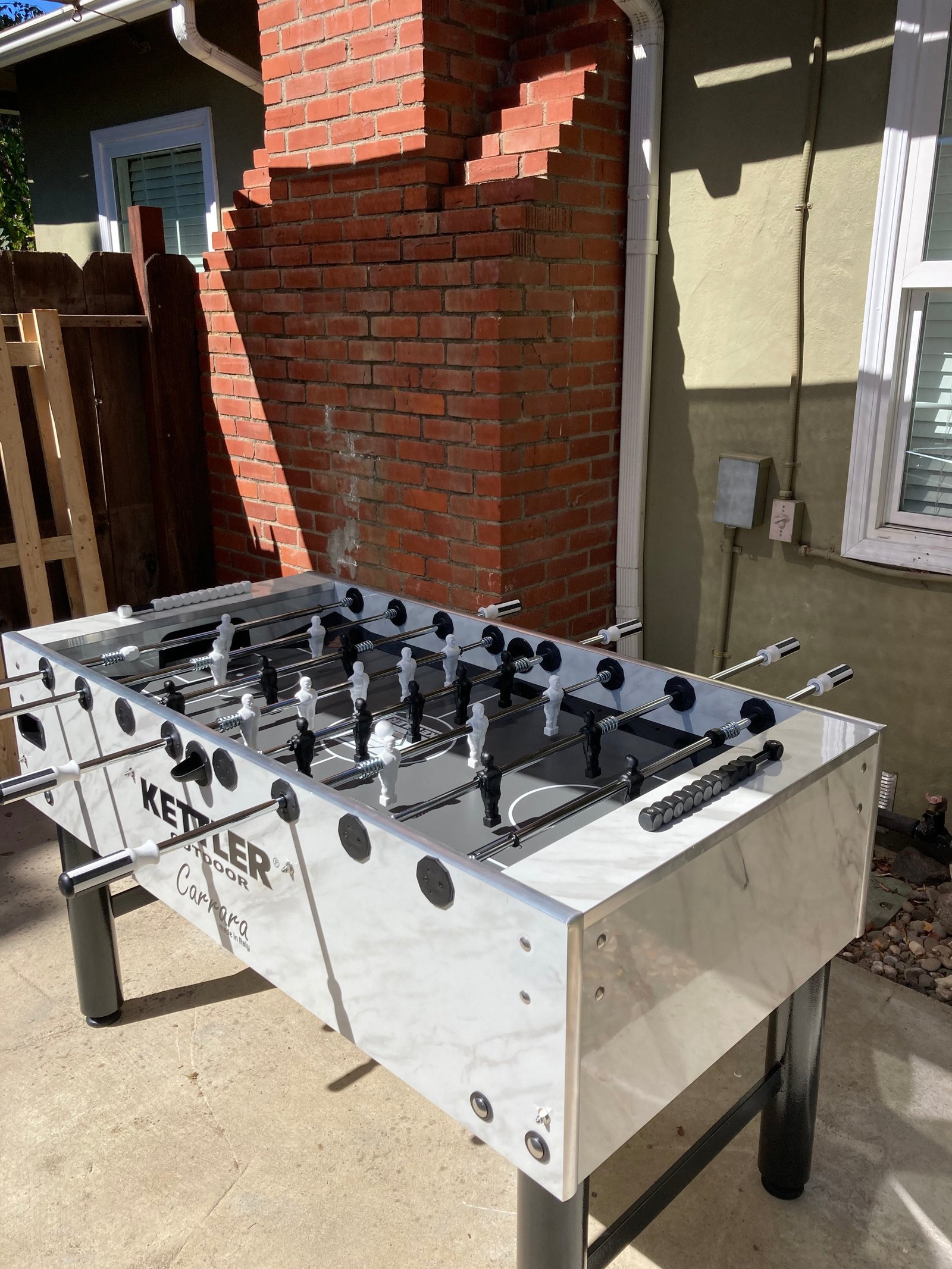 A Kettler Carrara weatherproof outdoor foosball table sits by a brick wall and window.