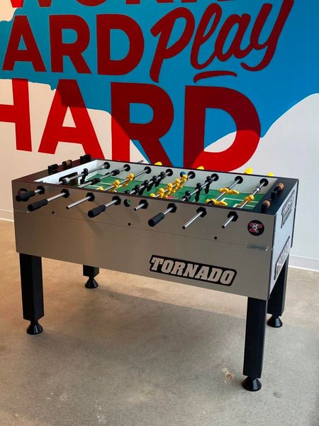 A Tornado Tournament Competition T-3000 Foosball Table in Silver by Tornado stands on a concrete floor, positioned in front of a wall that reads "Work Hard Play Hard" in blue and red letters.