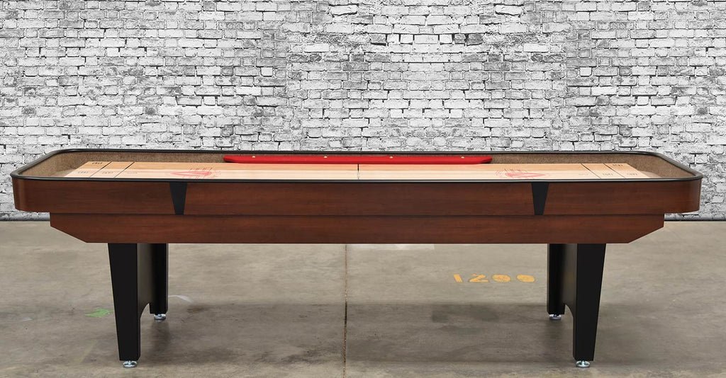 A Venture Classic Bank Shot 9' Shuffleboard Table with black legs stands on concrete by a brick wall.